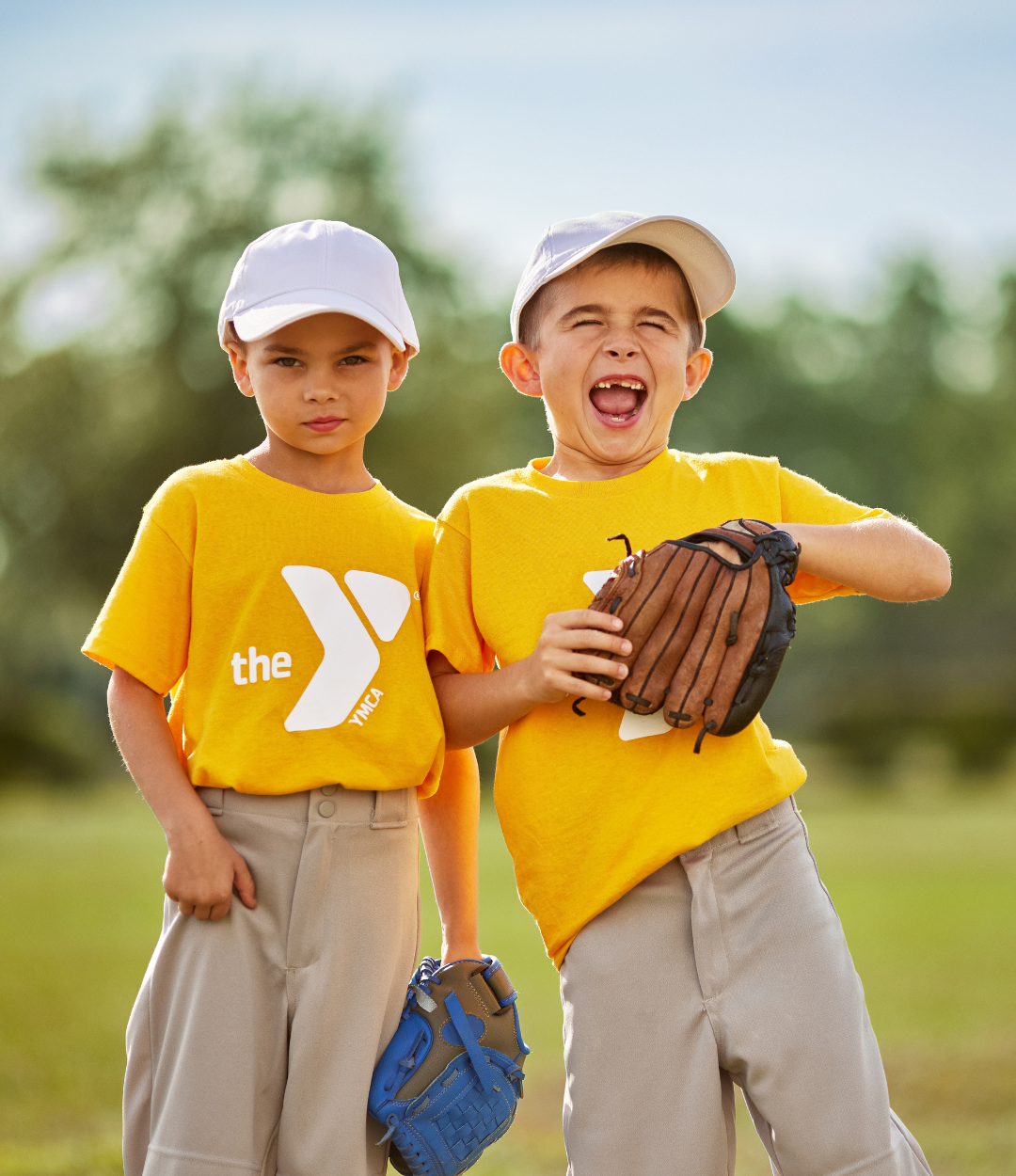 Two young boys smile holding their baseball gloves while outside at a YMCA baseball game.