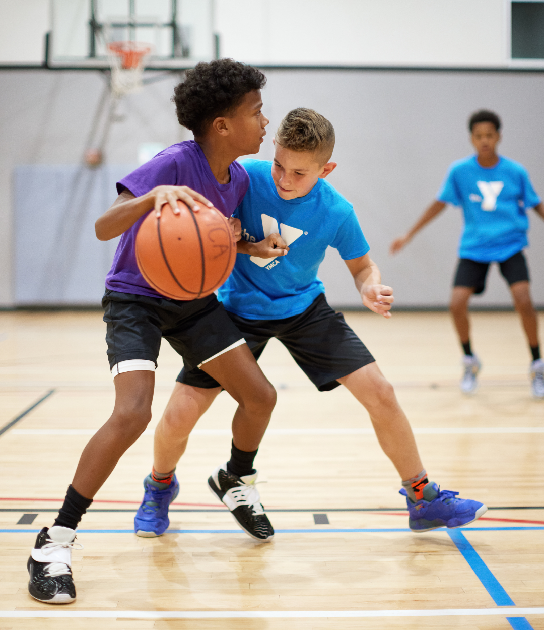 A teenage boy attempts to dribble a basketball closer to the hoop while another teenage boy on the opposing team tries to block him from getting closer at an indoor basketball game.